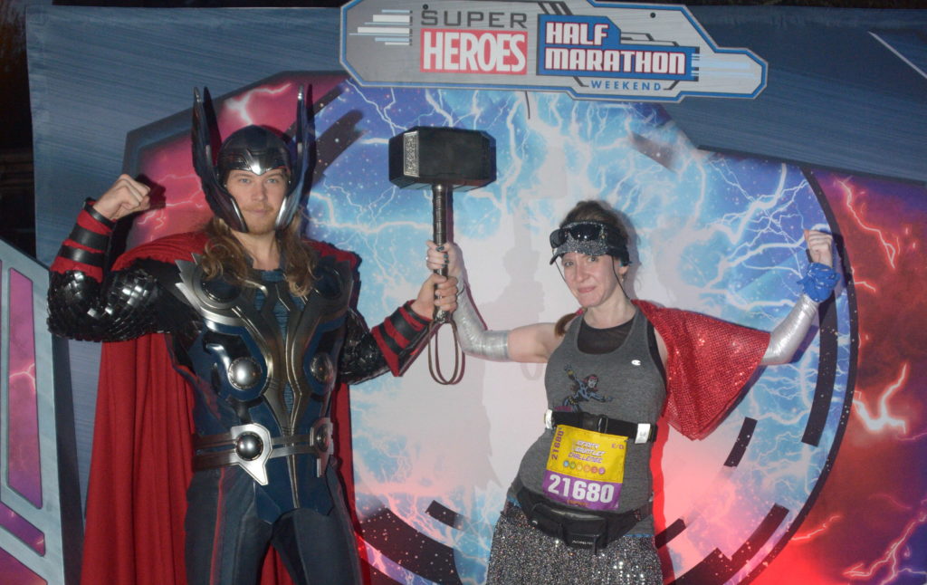 The author Cheryl Platz is wearing a race bib from a half marathon as she poses holding Mjolnir with an actor playing Thor at Disneyland.