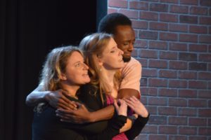 Cheryl performing in a scene at Seattle Theatresports, produced by Unexpected Productions in the Market Theater in Seattle's Pike Place Market. Cheryl is being hugged by fellow ensemble members Laurel Ryan and Issiah Harris in the midst of a musical scene.
