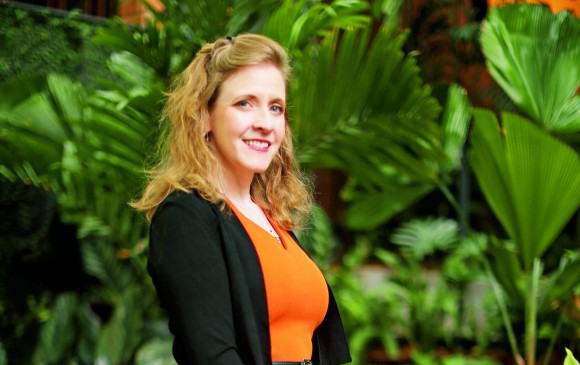 Cheryl Platz, a blonde woman with blue eyes wearing a black jacket and orange dress in front of a tropical background.