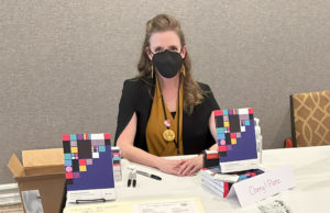 Cheryl sits at a book sales table at the Nebulas Conference 2023 in Anaheim with her book Design Beyond Devices on display.