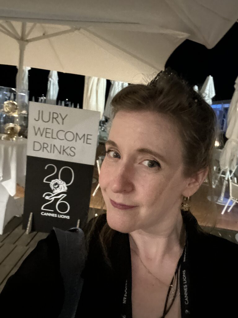 A selfie of Cheryl in front of the Jury Welcome Drinks sign at a beachfront venue in the evening.